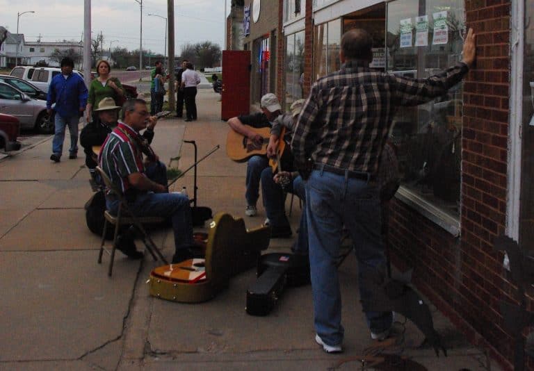 Two musicians play guitar while seated on the sidewalk outside a brick storefront during a community event, with pedestrians stopped to listen and watch
