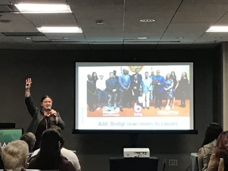 A woman with dark hair and beige skin tone wearing a businesslike jacket is speaking to a packed audience. She's holding a mic and raising one hand in the air. A slide shown on the screen behind her includes a diverse crowd at a golden shovel business event.