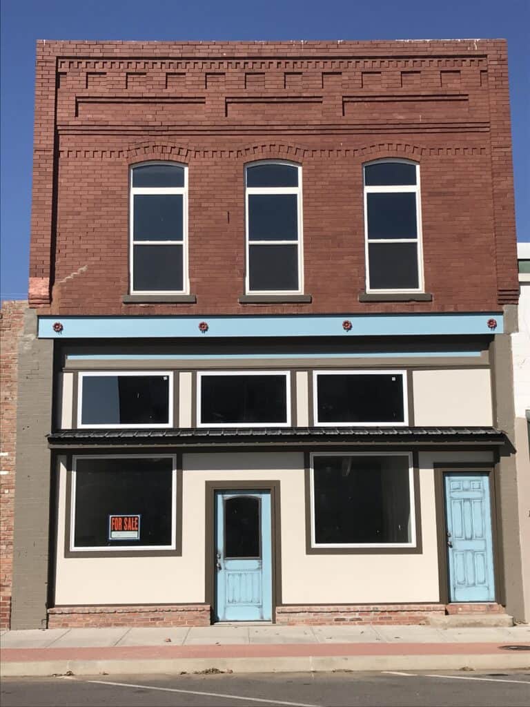 A two-story brick downtown building has been recently rehabbed with new windows and updated paint. A small "for sale" sign is in the window.