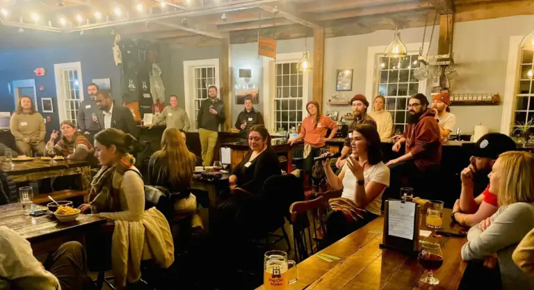 A group of diverse young people in a casual bar setting. One is holding a mic and speaking as others listen.