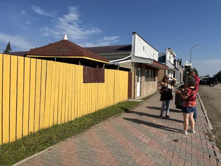 In a downtown, three people are standing on a sidewalk next to a yellow painted board fence. There are two small windows in the board fence that can open outward.