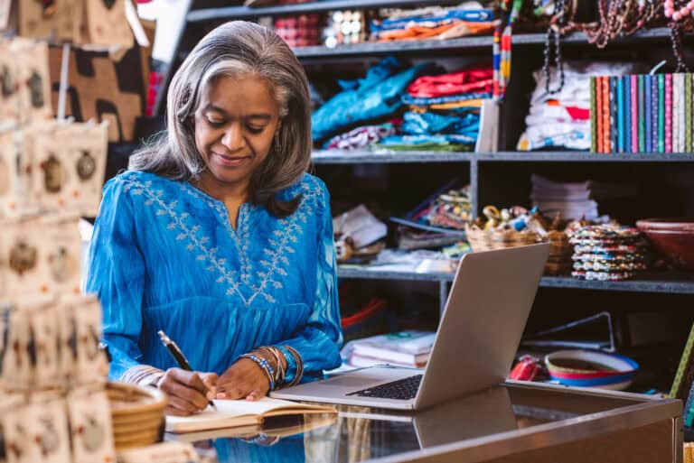 In a fabric store, a dark-skinned woman with long salt-and-pepper colored hair is using a laptop notebook to manage the business