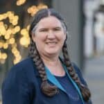 Becky McCray wearing long braids and a professional outfit smiles as she stands on a rural downtown street with twinkling lights in the background.