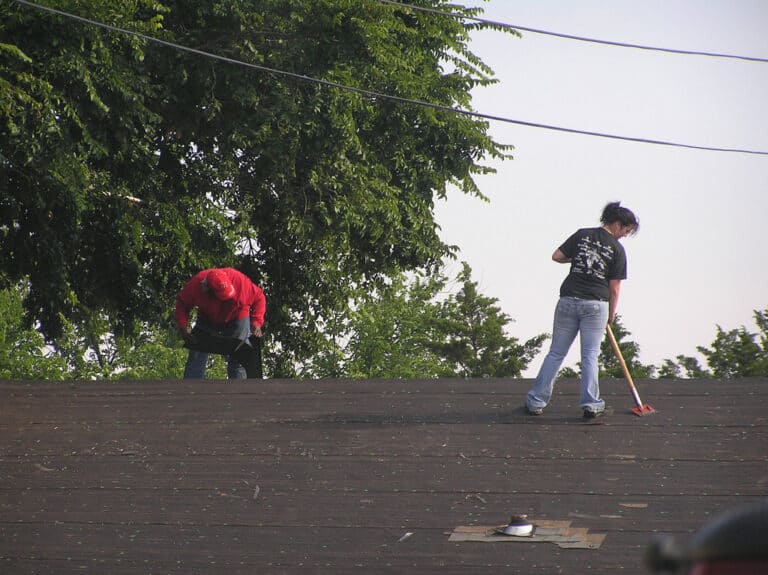 Two diverse workers installing new shingles on a roof. One worker in a red shirt cuts a shingle. A woman uses a push hoe to remove nails and debris.