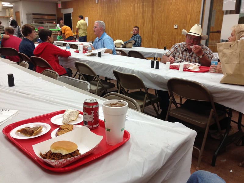 Red cafeteria tray with barbecue sandwich, beans, cookie, and drink sits on white-clothed table. Diverse diners of various ages eat and talk at long tables in community hall. Volunteers serve from kitchen area in background.