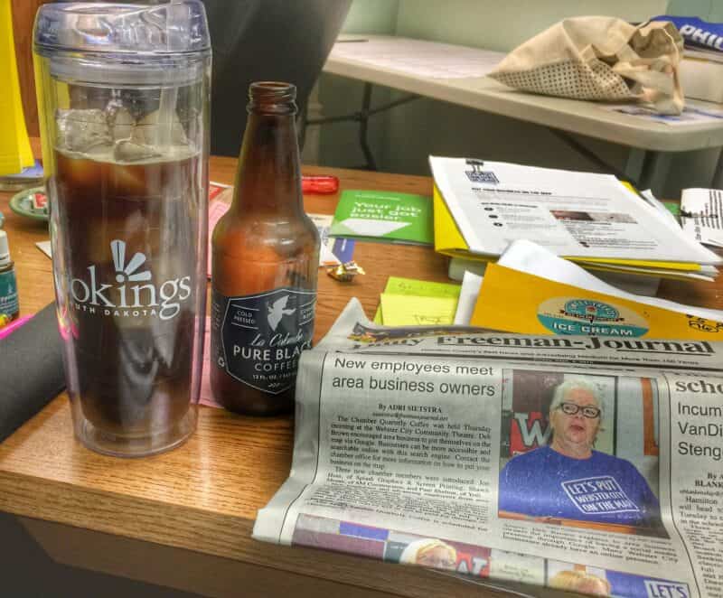 Clear coffee travel mug with iced coffee, and a local newspaper on a busy desk. 